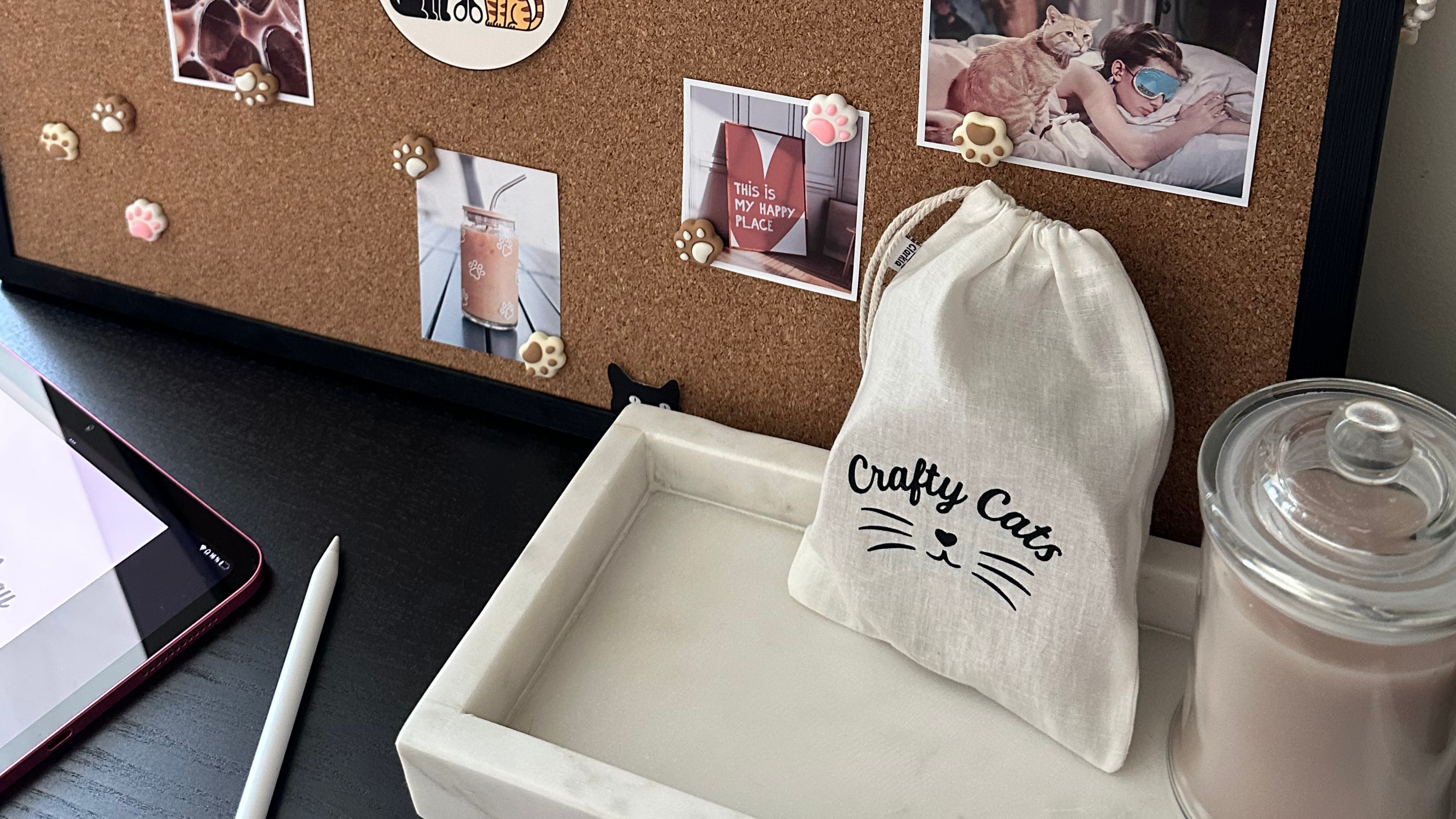 Cork bulletin board with photos, a drawstring bag labeled 'Crafty Cats', and a jar on a desk.